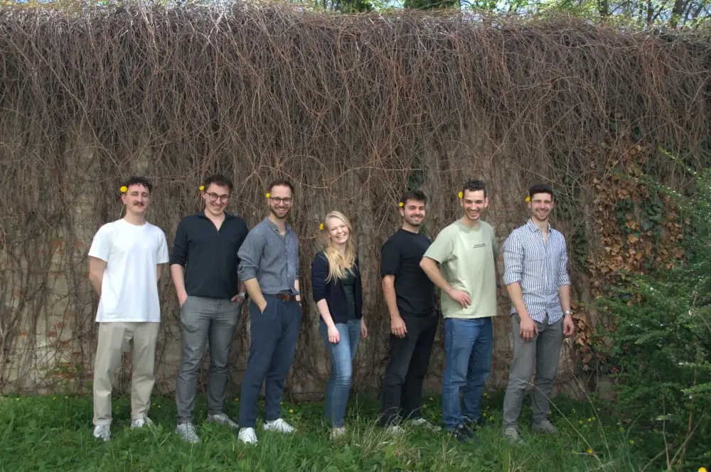 Sieben Teammitglieder stehen im Freien vor einer bewachsenen Wand, alle mit gelber Blume hinter dem Ohr